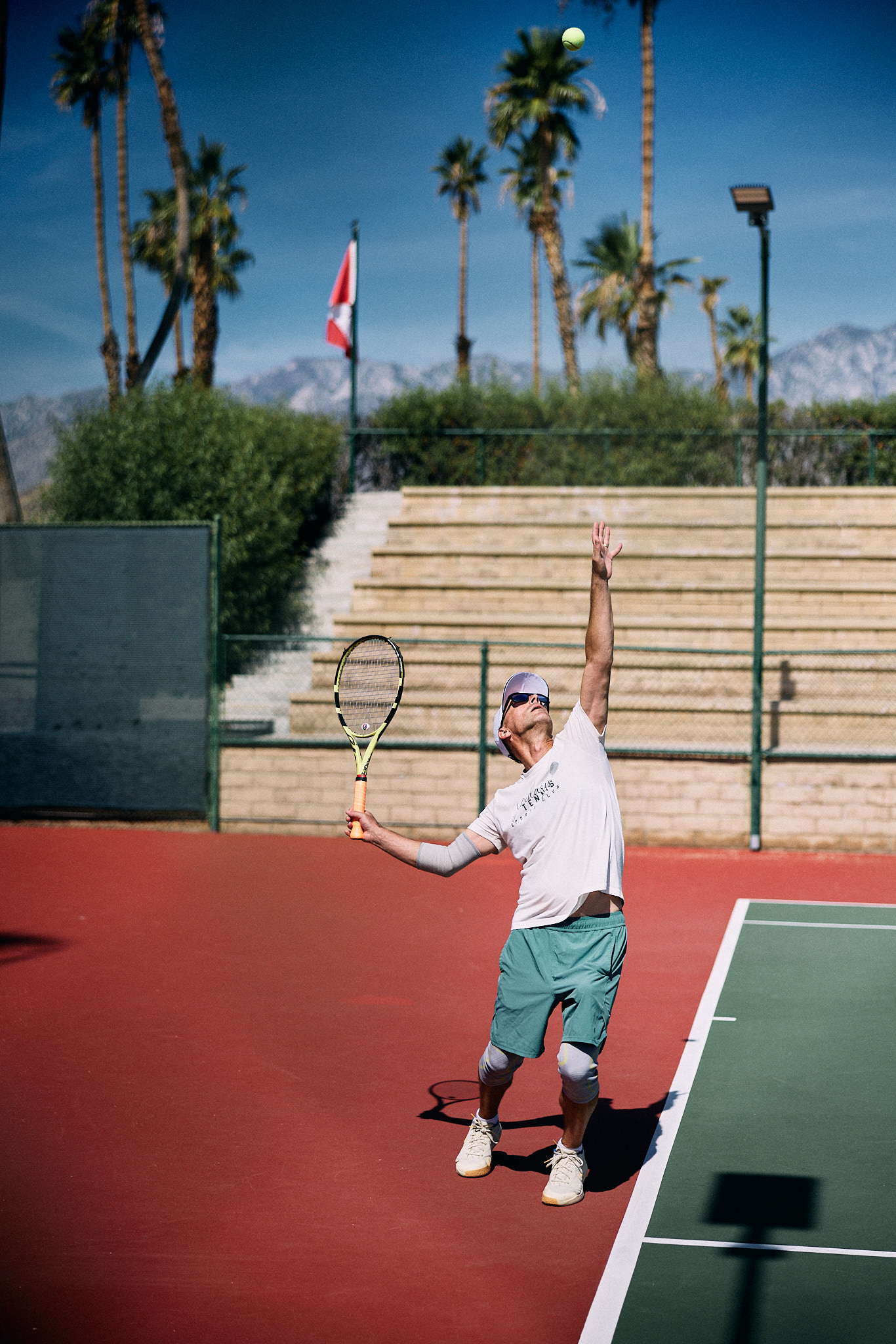 Coach Nathan Chesbro mid-serve, ball tossed high overhead, palms and the Santa Rosa Mountains behind the court at Cathedral Canyon.